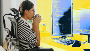 A woman drinking from a cup sitting in front of a desktop computer.