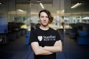 A man with an asymmetrical haircut, wearing a black T-shirt that says Stanford Medicine, stands with arms crossed in a glass office space