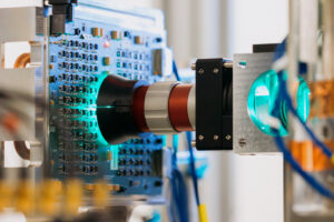 A close-up shows bluish light glowing through a lens that is surrounded by cables, wires and other computer hardware.