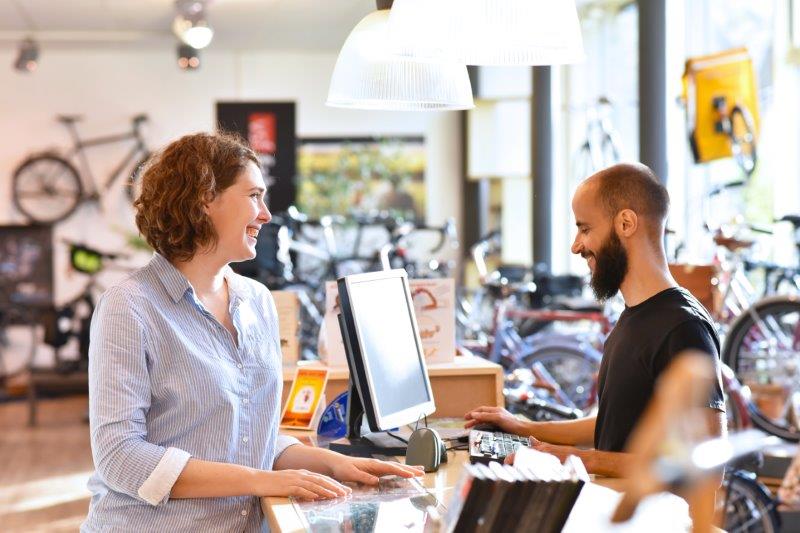Image of two people in a bike shop, one person behind a register, speaking to each other and smiling.