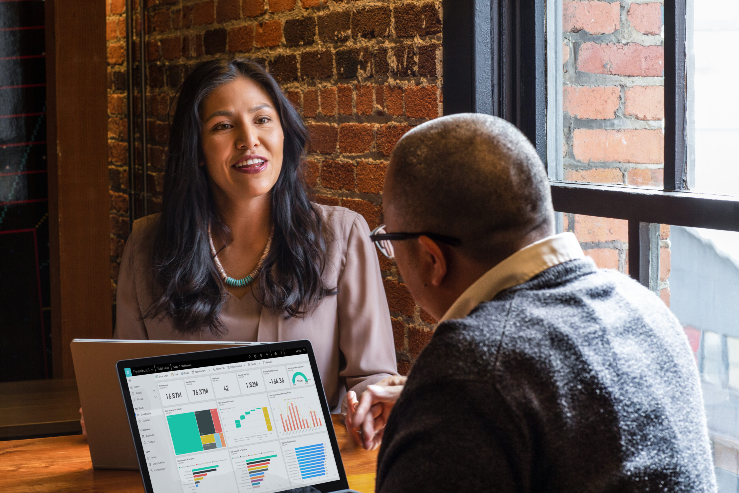a woman is talking to a man whose back is turned to the camera, they each have laptops in front of them.