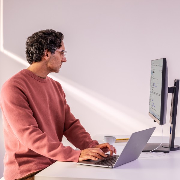 Man in focused work, standing at a desk working on a laptop and looking at a monitor.