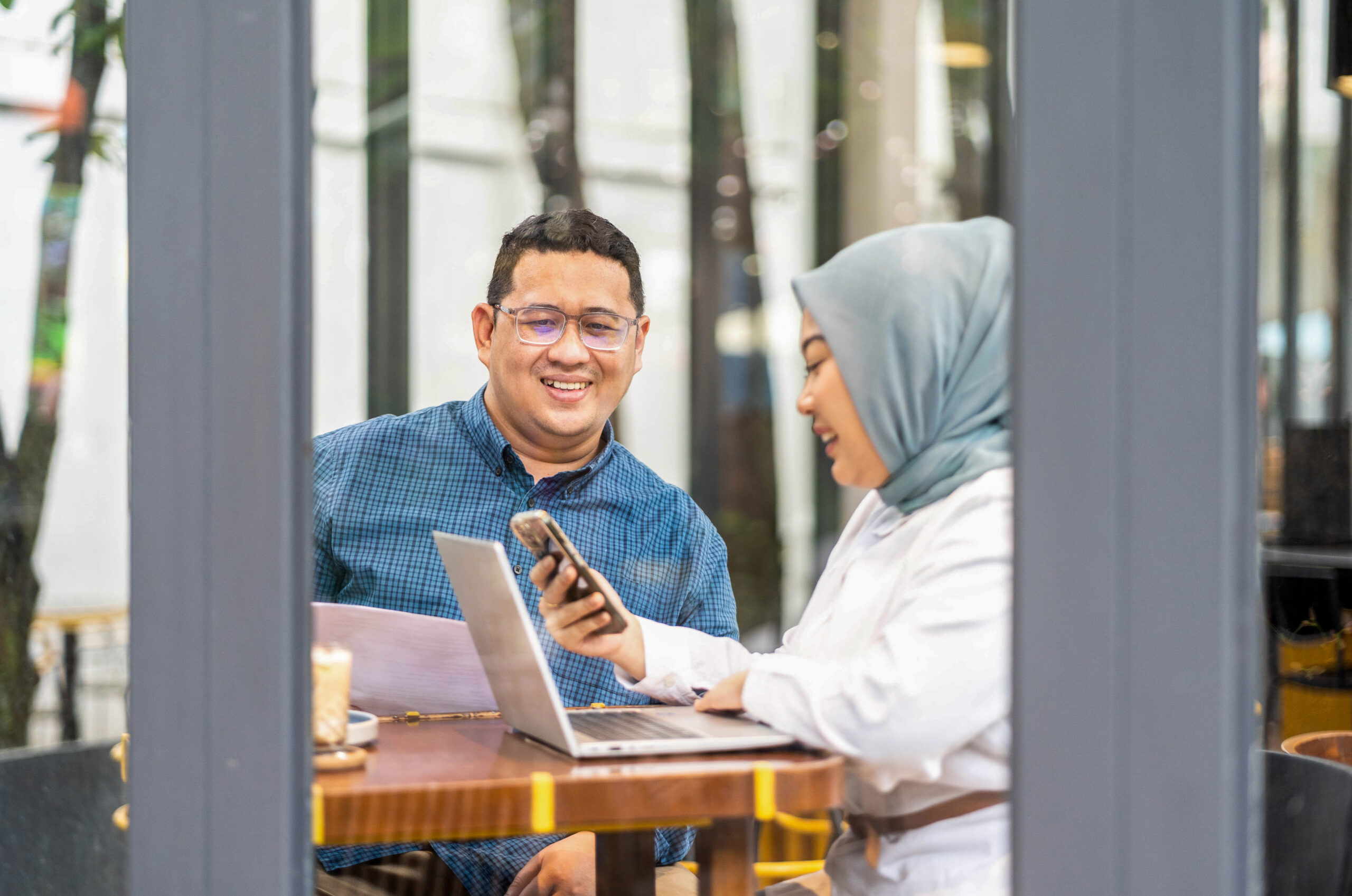 A man and woman sitting at a table looking at a laptop and a mobile device.
