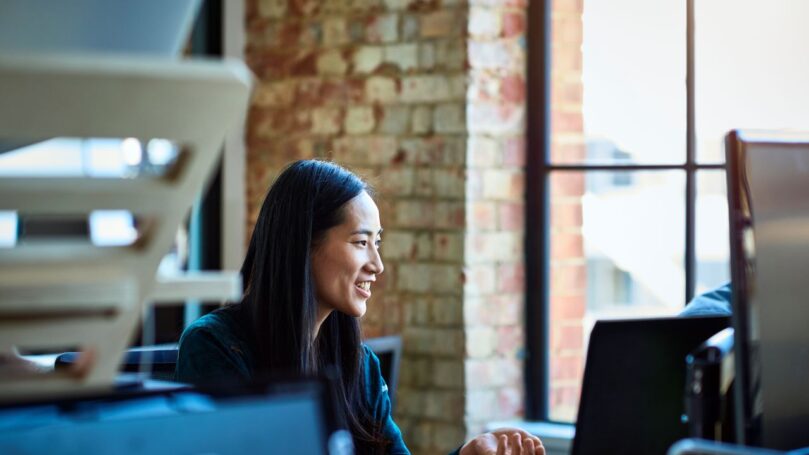Office worker, smiling and looking away, in contemporary studio with bare brick walls, sitting in front of a desktop computer.