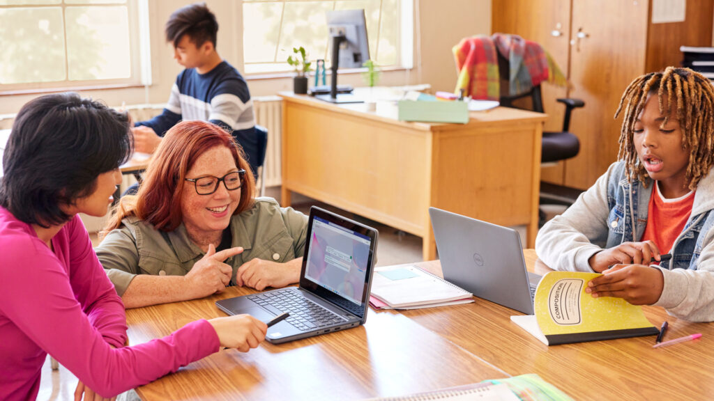 Early teen students working on laptops in a classroom while a teacher kneels by their table to engage with them.