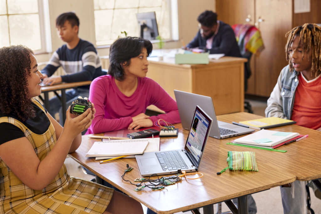 Three students sitting at a group of desks in a classroom working together on laptops. An additional student and a teacher are working individually in the background.