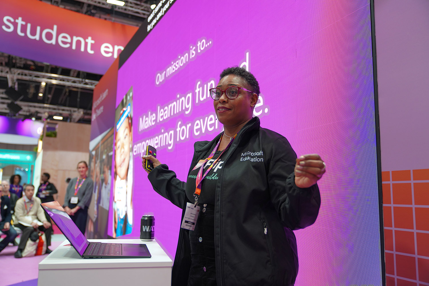 A presenter in a large conference hall standing in front of a large screen and speaking to attendees.