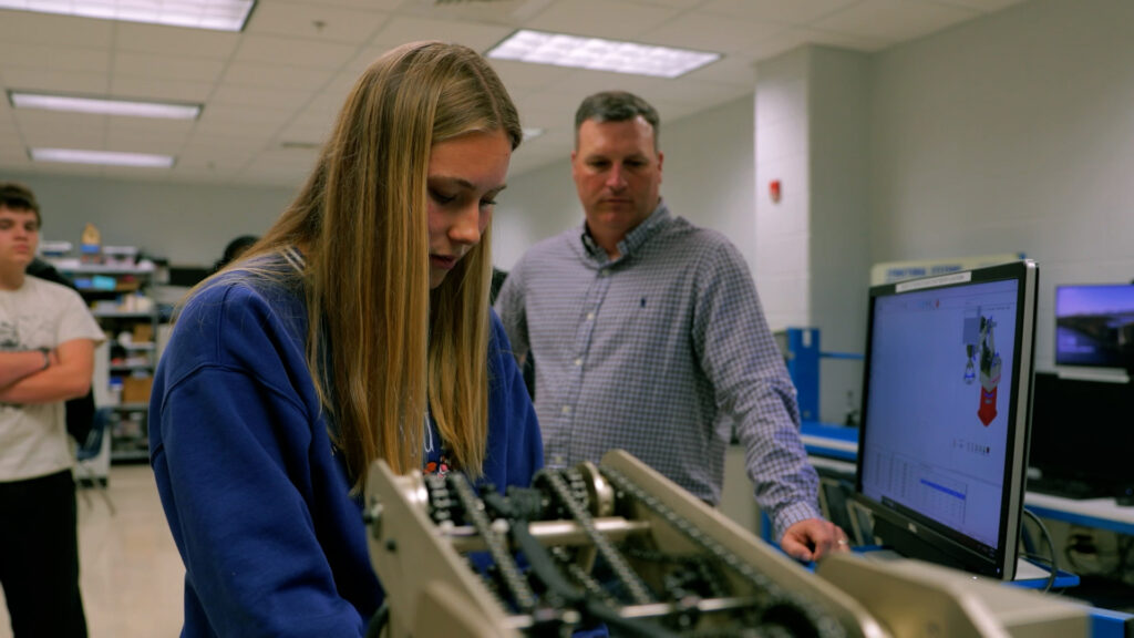 A technical classroom scene showing a student working on a robotic component from plans on a screen. A teacher and additional students look on from behind her.