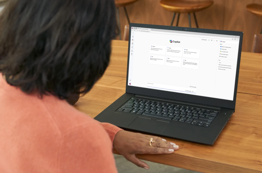 An educator sits at a table in a kitchen while using Microsoft 365 Copilot Chat on a laptop.