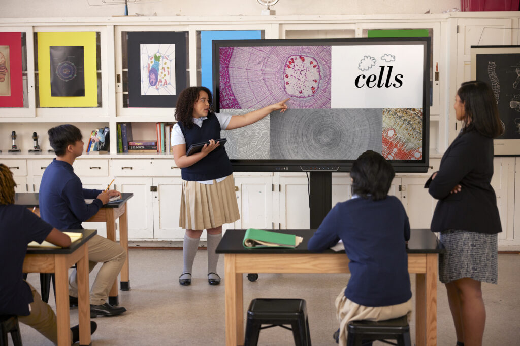 A uniformed elementary school student sharing a presentation on "cells" to a classroom of students and a teacher. 
