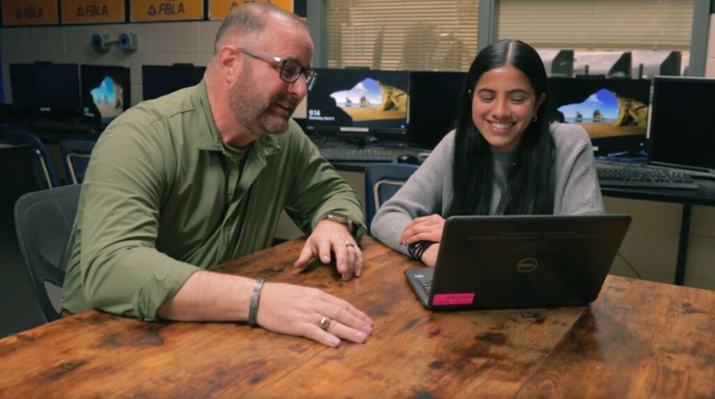 An educator and student sit together at a table in a classroom and look at the screen of a laptop.