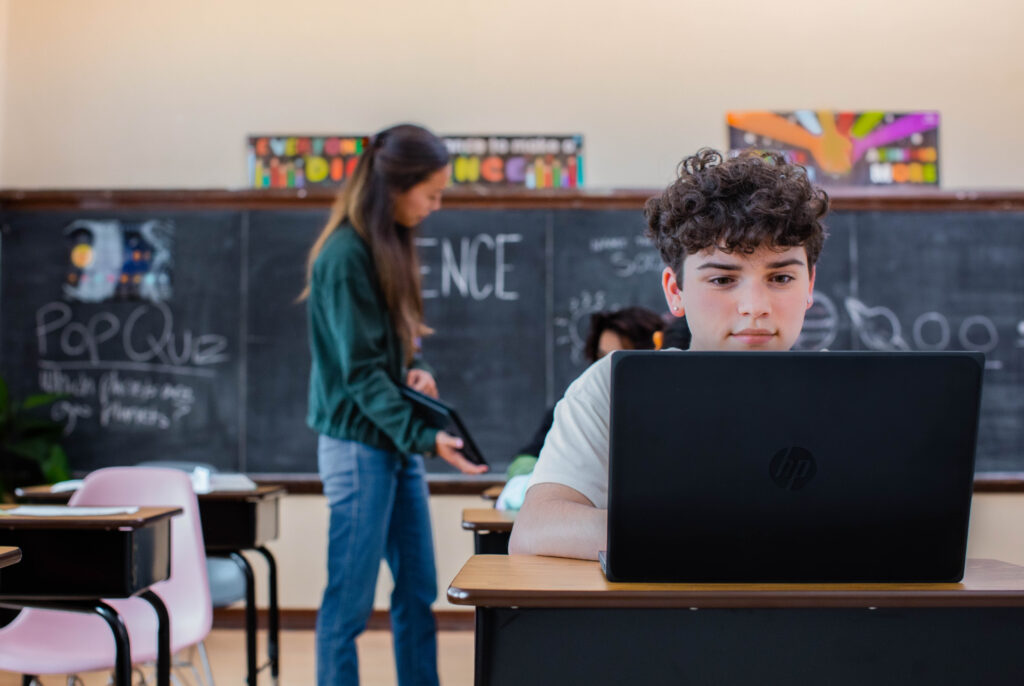 An elementary school student working on a laptop in a classroom setting while the teacher speaks to another student.