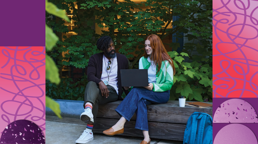 Two people sit together on a bench surrounded by trees and look at the screen of a laptop.