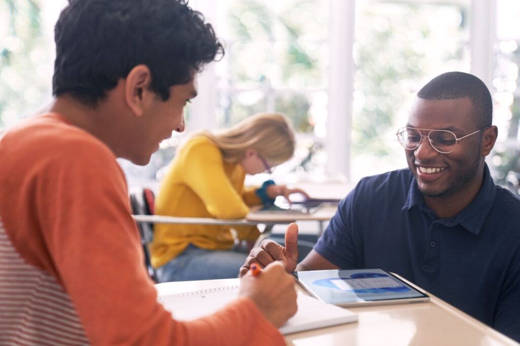 An educator kneeling at a student’s desk and helping him with schoolwork.