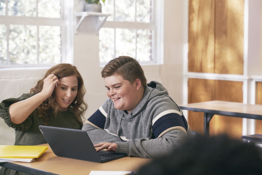 An educator kneeling at a student’s desk and helping him with schoolwork on his laptop.