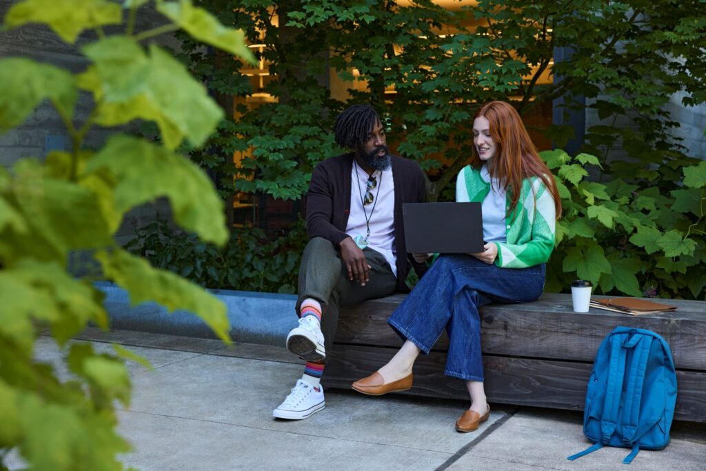 Two people in a higher education setting sit together on a bench surrounded by trees and look at the screen of a laptop.
