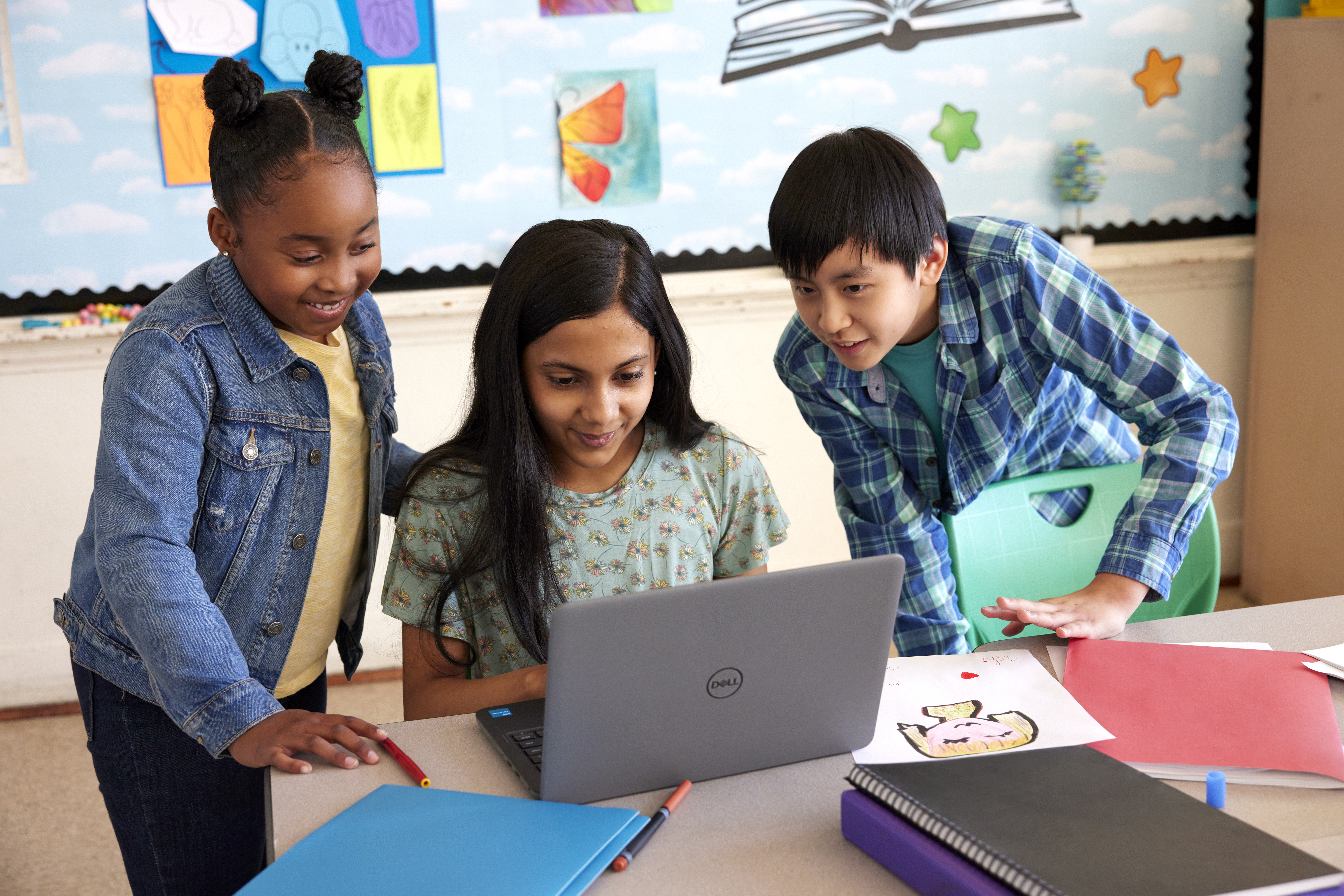 Three children in a classroom looking at a laptop together, engaged and focused on the screen.