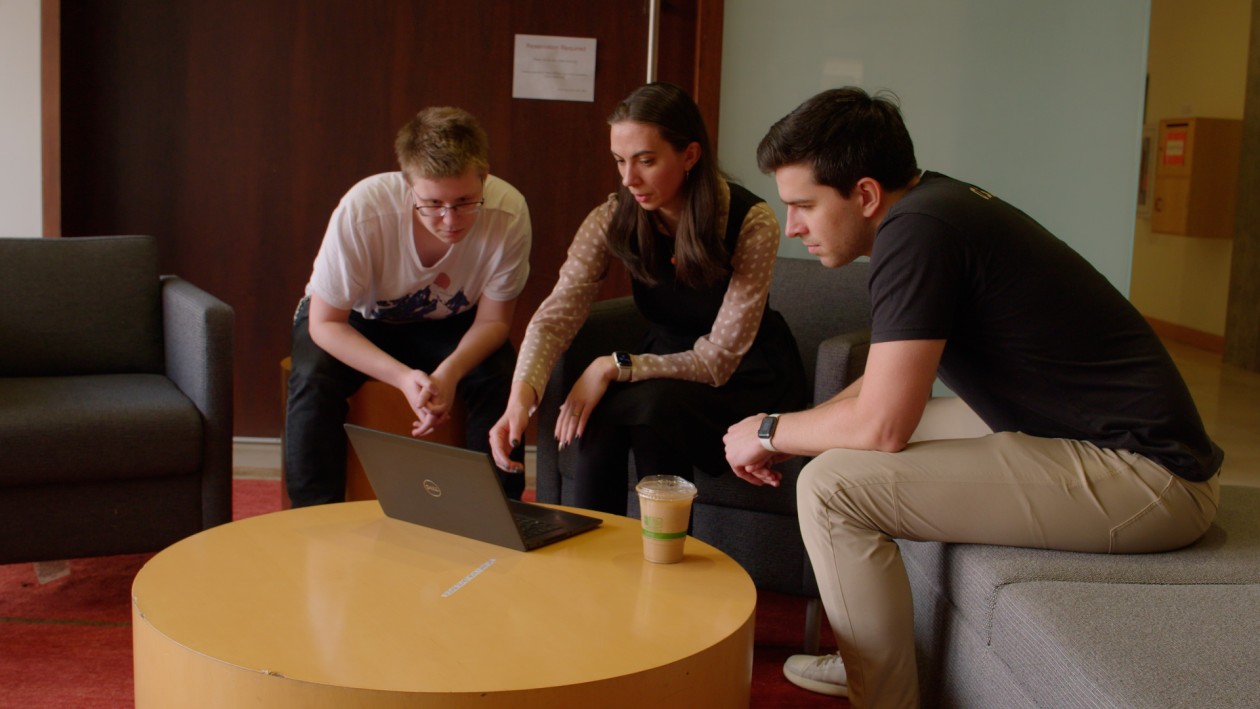 An IT professional and two students collaborating on a laptop outside of an office at a university.
