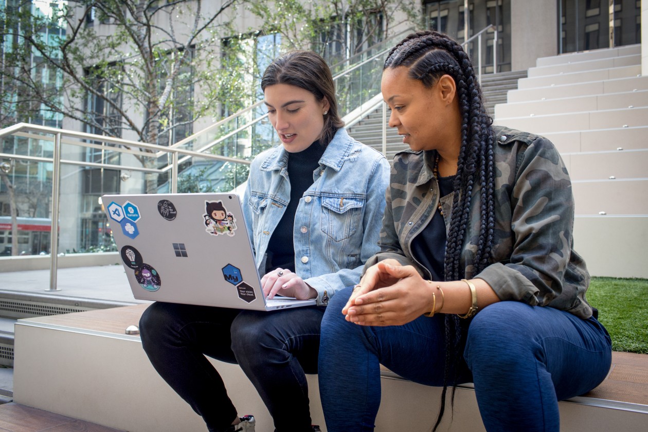 Two workers sit on the steps of an office building and work together on a laptop.