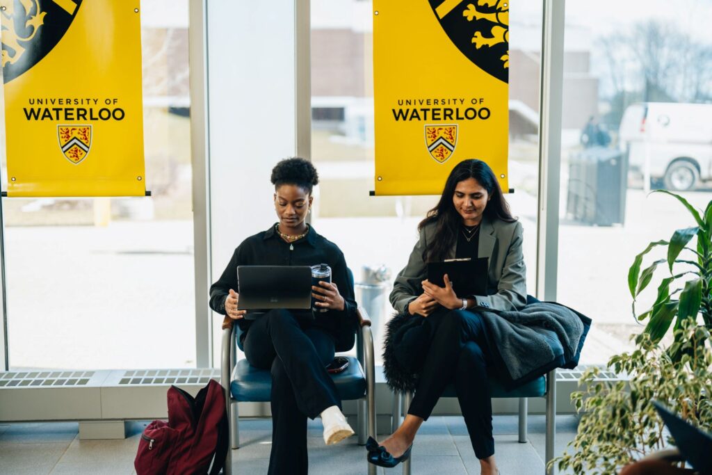 Two students sit below University of Waterloo banners while working on laptops.