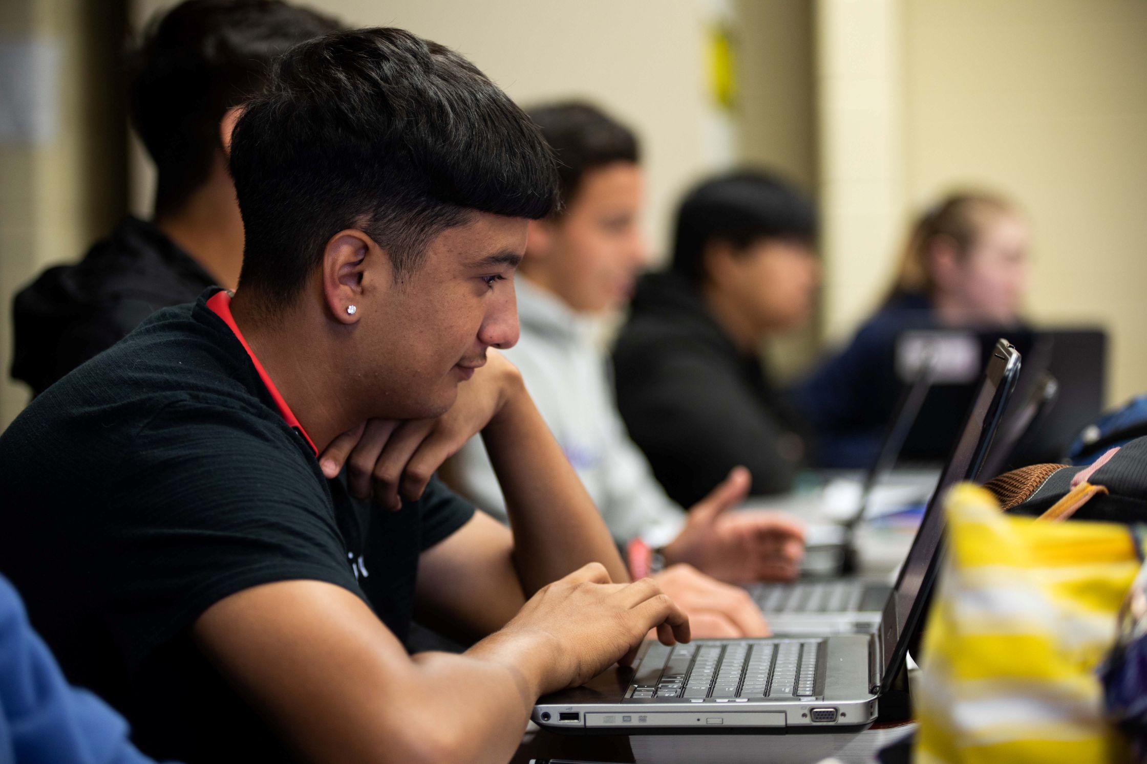 A classroom scene showing a row of students using laptops, focused on single student in the foreground.