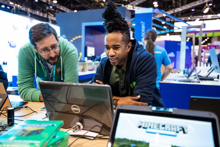 Two men work together on a laptop in a large conference hall.