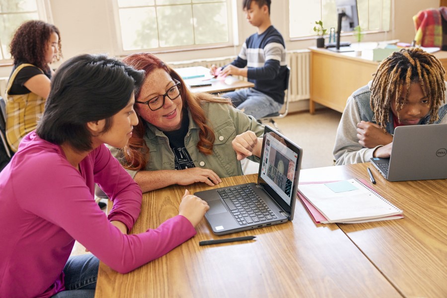 Decorative. A teacher kneeling near a student and they’re looking at the screen of a laptop together in a school classroom. There are other students nearby and in the background.