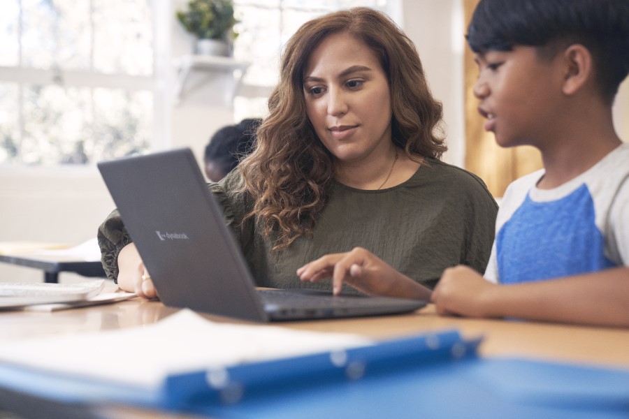 Decorative. A teacher engaging with a student who’s working on a laptop at a desk in a school classroom. 