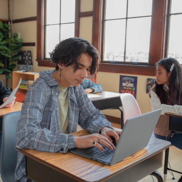 Decorative. A student uses a laptop in a classroom while other students work at their desks in the background.
