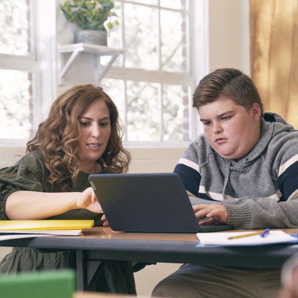 An educator kneeling at a student’s desk and helping him with schoolwork on his laptop.