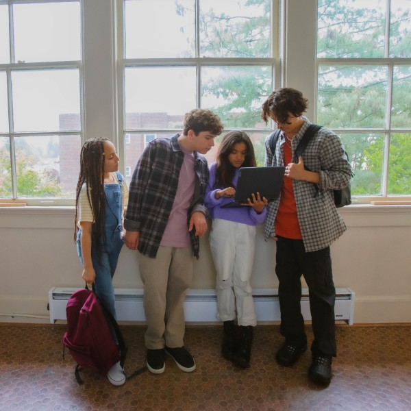 A group of students stand in a school hallway huddled together over a laptop.