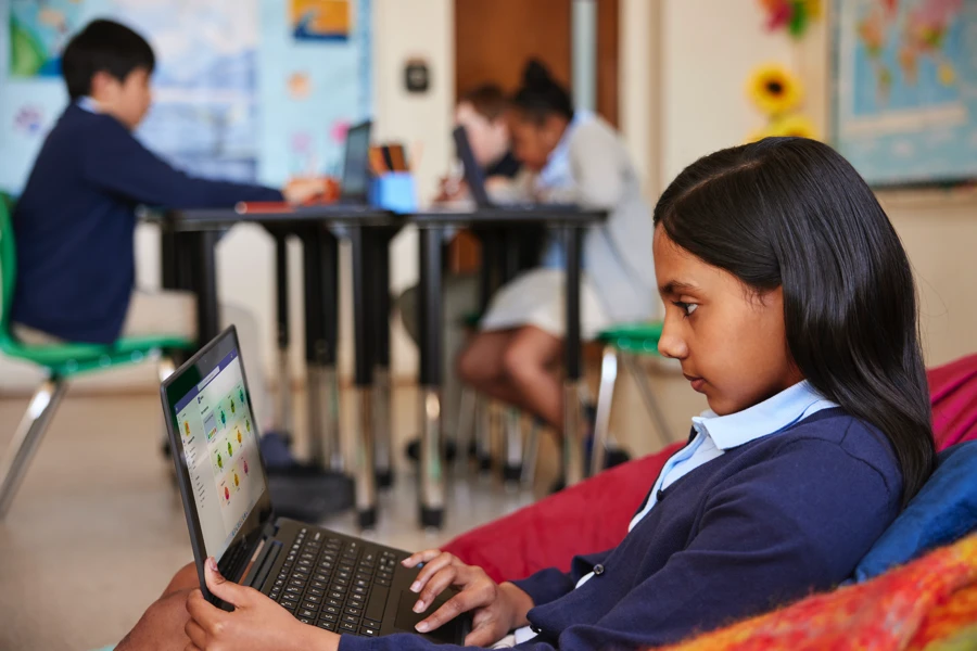 A student using a laptop sits on a beanbag in a classroom with students working at a table in the background.