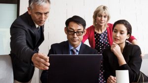 Four people sitting and standing at a desk analyzing a laptop computer