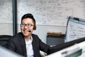 Male worker sitting at cubicle desk in financial office, wearing glasses and phone headset.