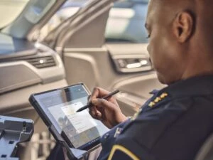 Male police officer holding a Surface go 3 in a ruggedized case with Teams calendar screen shown while sitting in a police car.