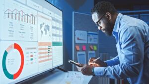 A man using a tablet and a stylus working in front of a large computer screen displaying graphs