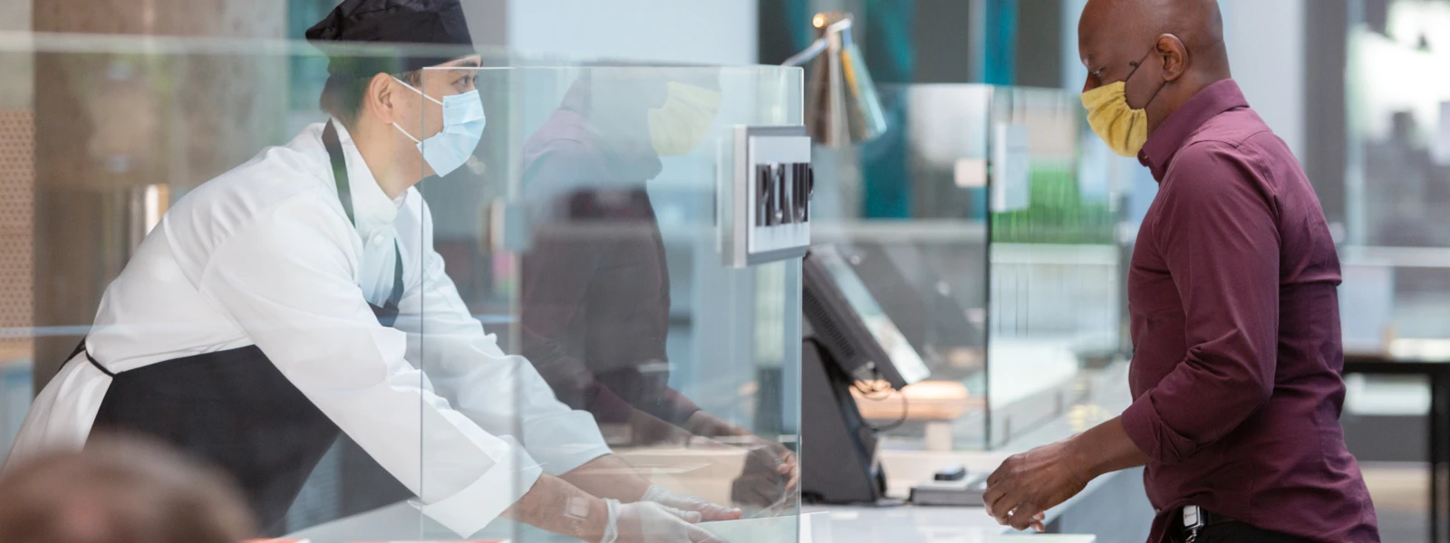 Ein Mitareiter im Lebensmitteleinzelhandel steht hinter einer Theke und gibt Essen aus. Er trägt Handschuhe und einen Mund-Nasen-Schutz.