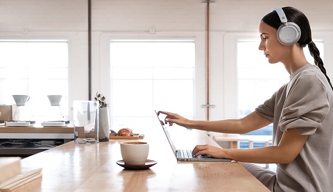 Eine Frau mit Kopfhörern sitzt in ihrer Küche und arbeitet an einem Surface Device.