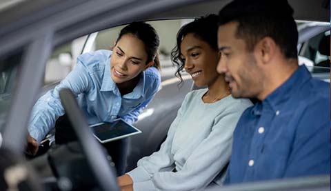 A man and a woman sitting in a car. Another woman is leaning in to explain something