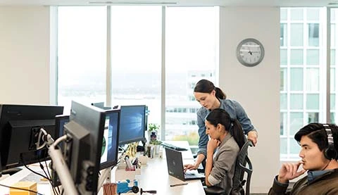 Drei Personen in einem B&uuml;ro, im Hintergrund gro&szlig;e Fenster. 