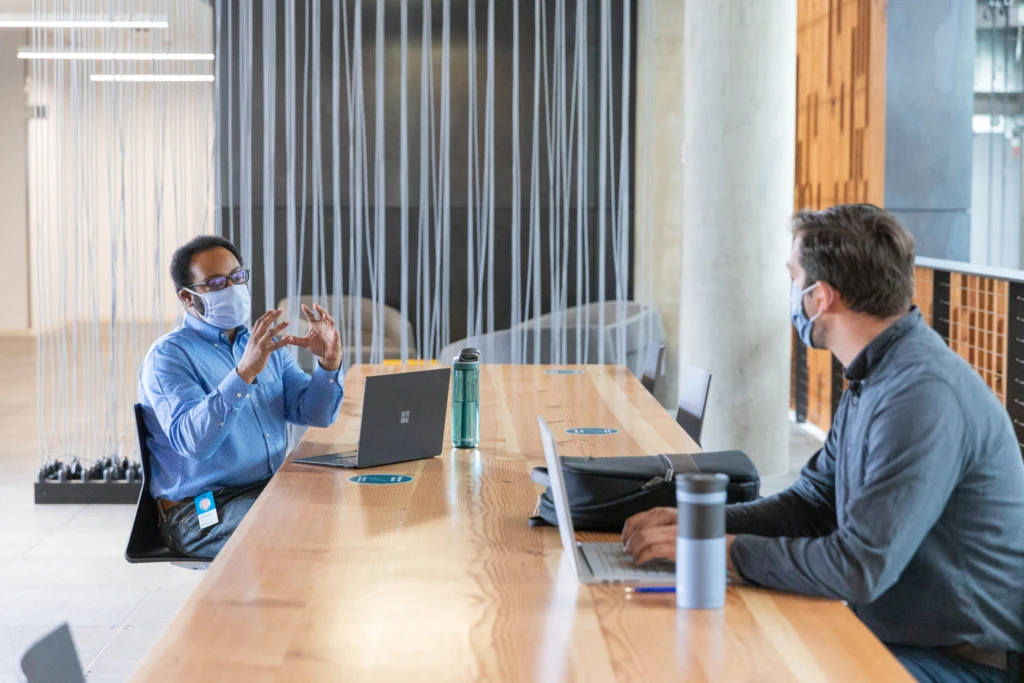 Two people in a meeting room, 2 metres apart wearing face masks. As part of a hybrid workplace, offices may be rejigged to focus on collaboration and meeting spaces.