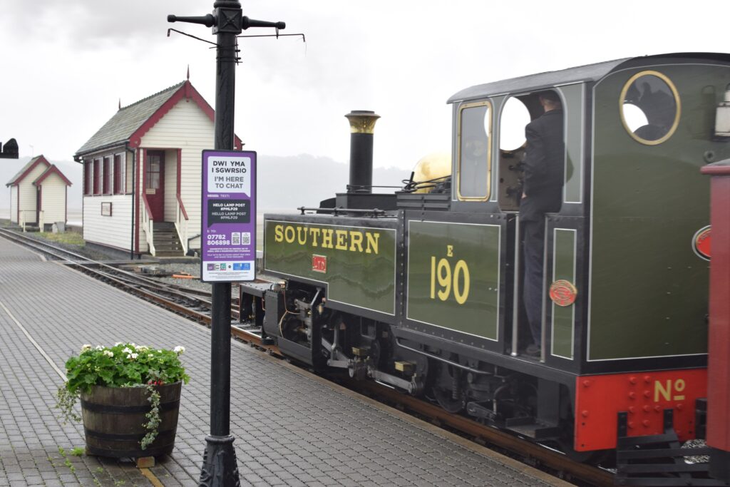 A Hello Lamp Post sign on a lamp post in Porthmadog, Wales. The sign is located on the platform of a train station.