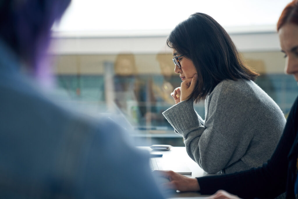 Worker with glasses sitting at a community table in common area reading on a laptop with other devices nearby.  