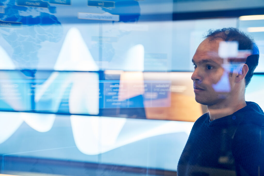 IT team member inside a secure room looking at data displayed on a large monitor behind glass walls with reflections.