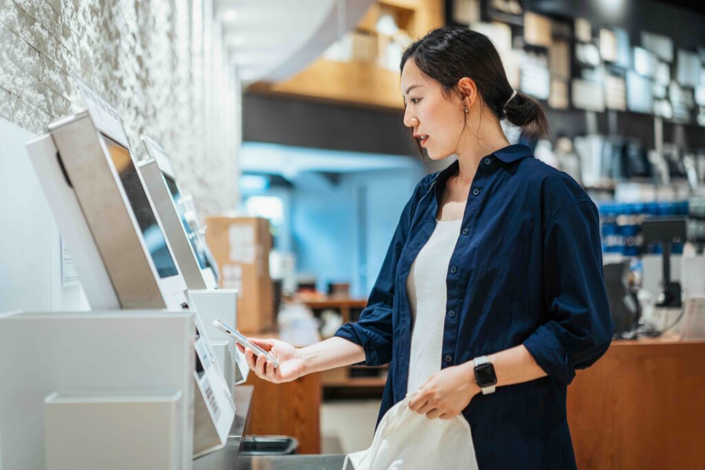 Young Asian woman with a reusable shopping bag, using contactless payment via smartphone to pay for her shopping at self-checkout kiosk in a store