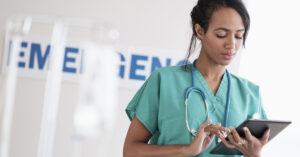 Healthcare professional in teal scrubs using a tablet, with a stethoscope around their neck, standing in a clinical setting with medical equipment in the background.