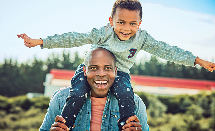 A man with a small boy on his shoulders walks outdoors. The two are smiling, and the child has his arms out like an airplane.