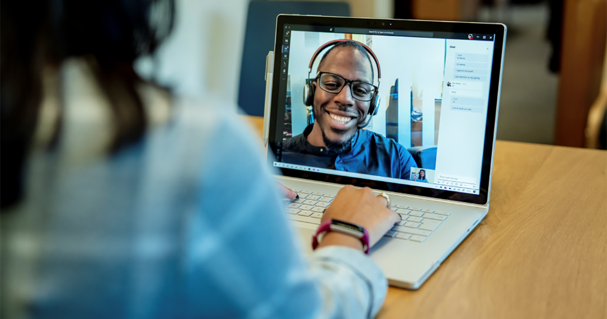 Image of a woman at a desk using a Surface laptop to make a Microsoft Teams video call with one man smiling and wearing a headset.