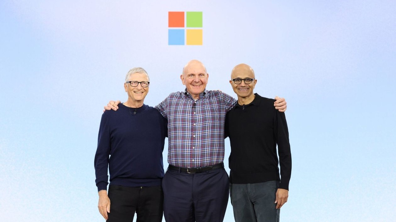 Three men with their arms around each other&rsquo;s shoulders stand on stage in front of the colorful square Microsoft logo.