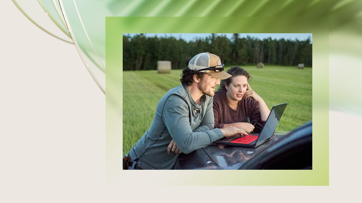 Two people looking at a laptop on the hood of a car parked in a hayfield.